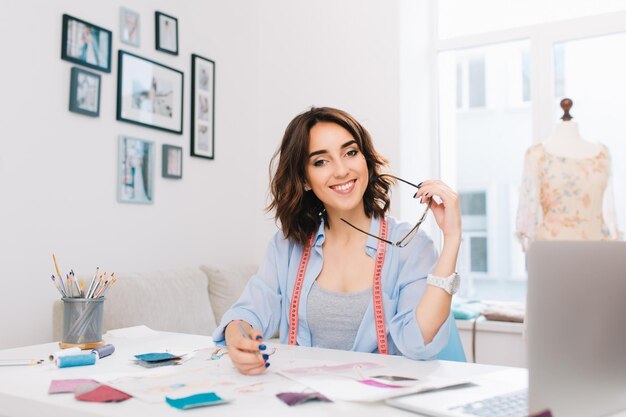 A brunette girl is sitting at the table in the workshop. She has blue shirt and creative mess on the table. She holds glasses and pencil in hands and smiling to the camera.