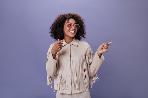 Brunette girl in lilac glasses and beige outfit shows her finger to side Happy woman in stylish jacket smiling on purple backdrop