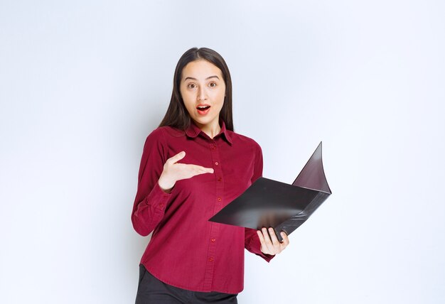 A brunette girl model standing and posing with a folder against white wall.