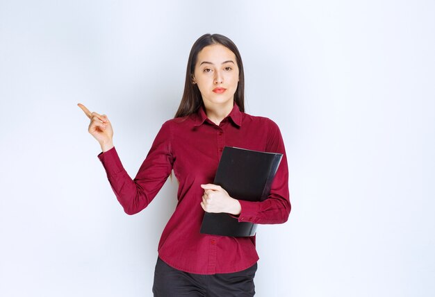 Free photo a brunette girl model standing with a folder and pointing away with an index finger.