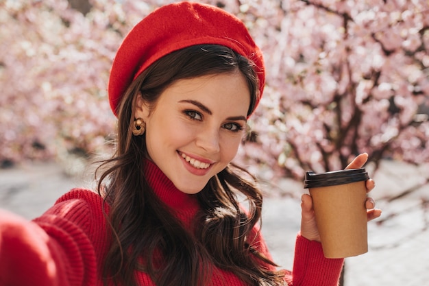 Foto gratuita ragazza bruna in berretto rosso fa selfie con un bicchiere di caffè. donna dagli occhi verdi in maglione di cachemire ampiamente sorridente e tenendo la tazza di tè