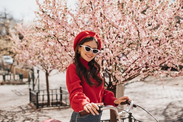 Brunette girl in red hat and sweater posing against background of sakura. Charming woman is stylish sunglasses smiling and riding bicycle