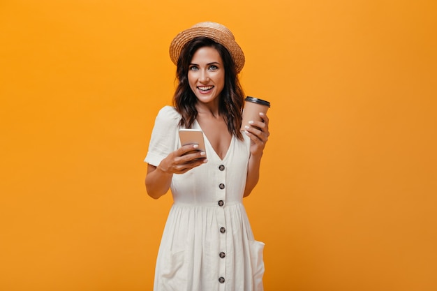 Brunette girl in straw hat holding smartphone and glass of coffee. Woman with wavy hair looks at camera with phone and with glass of tea in her hands.