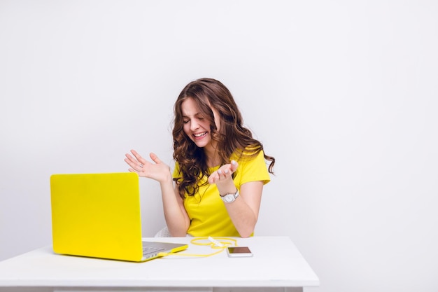 곱슬머리를 한 갈색 머리 소녀가 노란색 케이스에 노트북 앞에서 웃고 있습니다. 그녀는 흰색 테이블 뒤에 앉아 노란색 티셔츠를 입고 있습니다. 테이블에 노란색 충전기에 스마트폰이 있습니다.