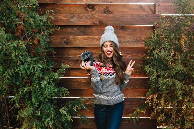 Brunette girl with long hair in winter clothes having fun with camera in hands on wooden  outdoor surround green branches.