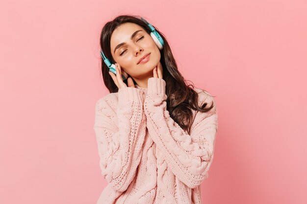 Brunette girl with pleasure listens to music on headphones. Woman in pink outfit smiling on isolated background.