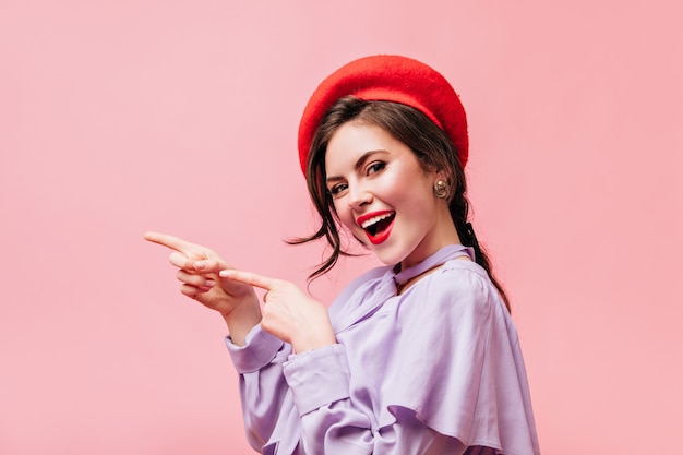 Brunette girl with red lipstick is smiling and pointing her fingers to left. portrait of woman in beret with place for text on pink background.