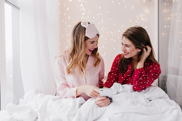 Brunette girl with shy smile showing phone to sister while chilling in bed. Indoor portrait of pleasant female friends relaxing in weekend morning.
