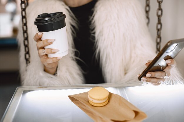 Foto gratuita ragazza bruna. donna in una pelliccia bianca. signora con telefono e caffè.