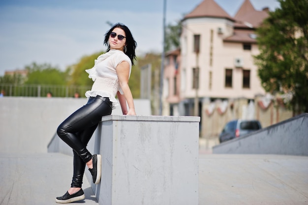 Brunette girl on women's leather pants and white blouse sunglasses posed against skate park
