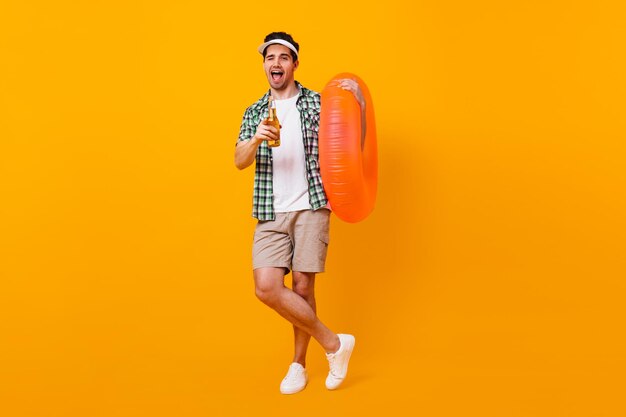 Brunette guy in cap winks and poses with inflatable circle Fulllength portrait of man in shorts with bottle of beer