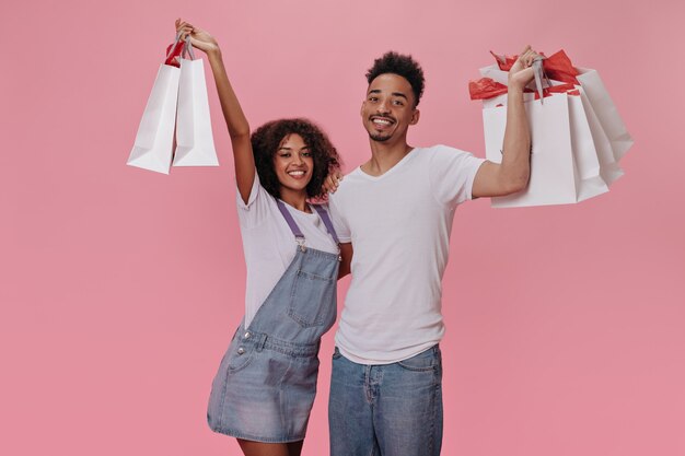 Brunette guy and girl happily posing with shopping bags on pink wall