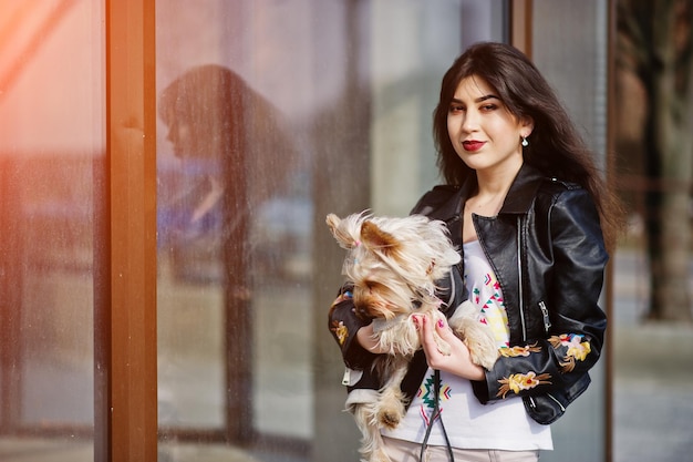 Brunette gypsy girl with yorkshire terrier dog posed against large windows house Model wear on leather jacket and tshirt with ornament pants
