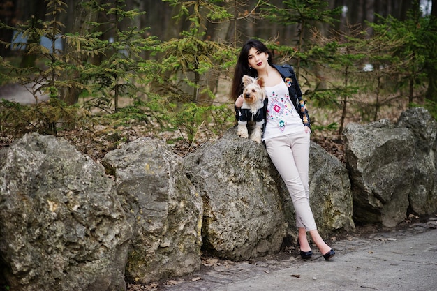 Brunette gypsy girl with yorkshire terrier dog posed against stones on park Model wear on leather jacket and tshirt with ornament pants and shoes with high heels