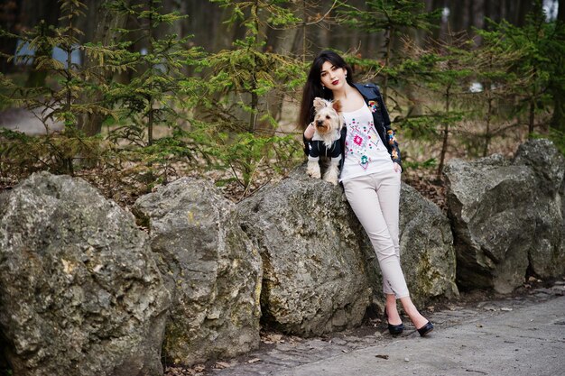 Brunette gypsy girl with yorkshire terrier dog posed against stones on park Model wear on leather jacket and tshirt with ornament pants and shoes with high heels