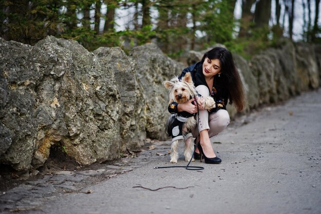 公園の石に対してポーズをとったヨークシャーテリア犬とブルネットのジプシーの女の子革のジャケットと装飾パンツとハイヒールの靴とTシャツのモデルの着用