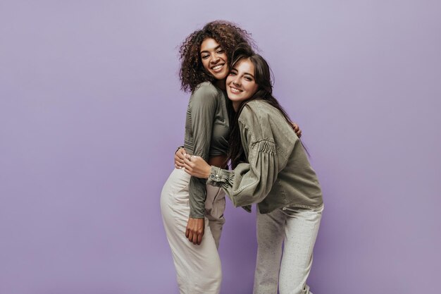 Brunette haired woman in stylish jacket smiling and hugging with stylish girl in olive top and light trousers on lilac background