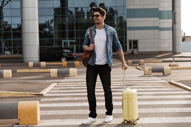 Brunette handsome man in white tshirt and black pants walks on crosswalk Cool guy in denim jacket and sunglasses holds brown backpack and yellow suitcase