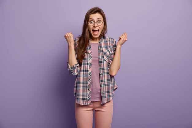 Brunette happy girl clenches fists like winner, has surprised overjoyed facial expression, keeps mouth opened, wears stylish checkered shirt, poses and gestures over purple wall, got victory