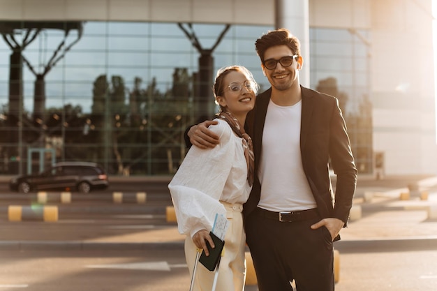 Brunette happy man in white shirt and black suit hugs attractive girlfriend near airport Charming lady in white blouse and eyeglasses smiles and holds passport