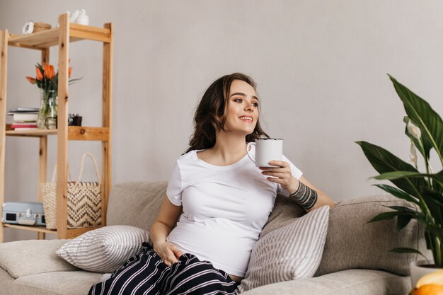 Brunette happy pregnant woman in white tee and striped pants gently smiles and holds cup of tea