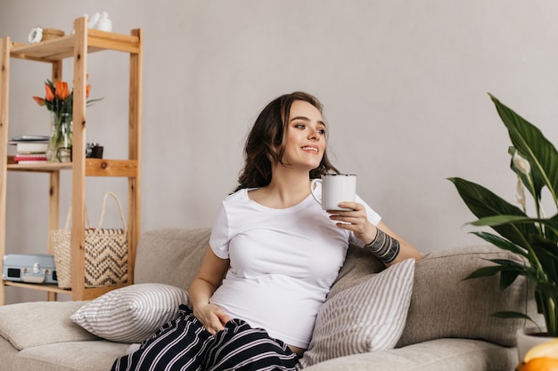 흰색 티와 줄무늬 바지에 갈색 머리 행복 임신 한 여자는 부드럽게 미소 짓고 차 한잔을 보유하고 있습니다.