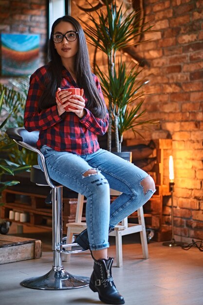 A brunette hipster female dressed in a fleece shirt and jeans drinks coffee in a room with loft interior.