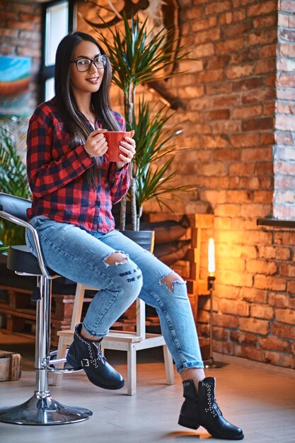 A brunette hipster female dressed in a fleece shirt and jeans drinks coffee in a room with loft interior.