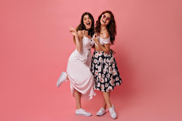 Brunette lady shows peace sign and poses with her friend on pink background Girls with wavy hair in white fashionable dresses and converse hugging