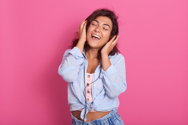 Brunette lady wears blue shirt, jeans and pink top, listening to music in headphones