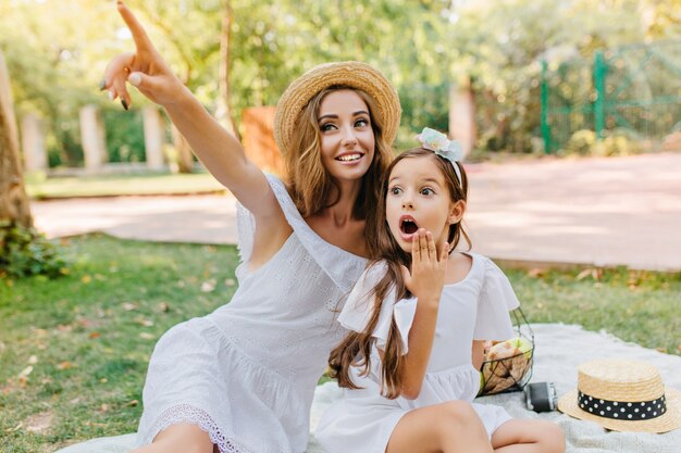 Brunette long-haired kid posing with shocked face expression on nature. Stunning young woman in white attire and vintage hat looks at something interesting and pointing with finger.