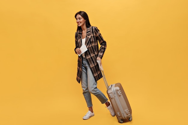 Free photo brunette longhaired woman in denim pants and brown coat poses with tickets and suitbag portrait of happy travel girl moves and smiles on isolated yellow background