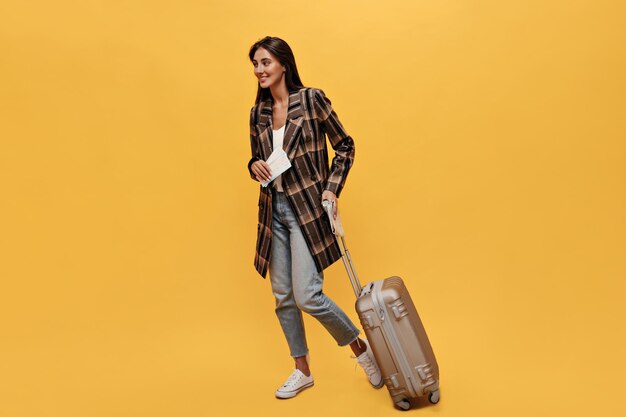 Brunette longhaired woman in denim pants and brown coat poses with tickets and suitbag Portrait of happy travel girl moves and smiles on isolated yellow background