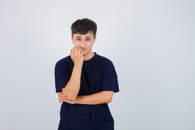 Brunette man biting him nails in t-shirt and looking thoughtful , front view.