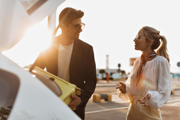 Brunette man in black jacket and blonde woman in white blouse smile and talk outside Handsome guy in sunglasses puts yellow suitcase into car trunk