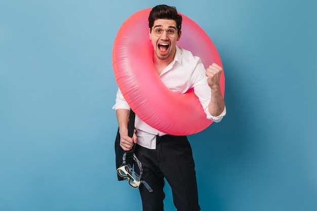 Free photo brunette man in black trousers and white shirt emotionally and joyfully screams. guy in glasses holds black bag, mask for swimming and puts on inflatable circle.