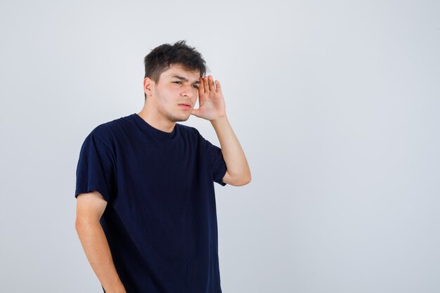 Brunette man in dark t-shirt holding hand to see clearly and looking unsure .