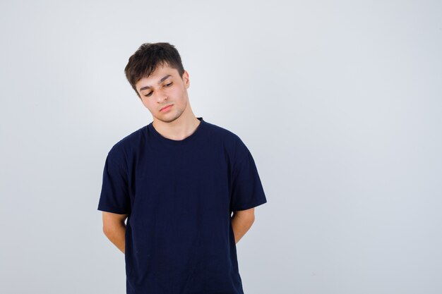 Brunette man holding hands behind back in t-shirt and looking pensive , front view.