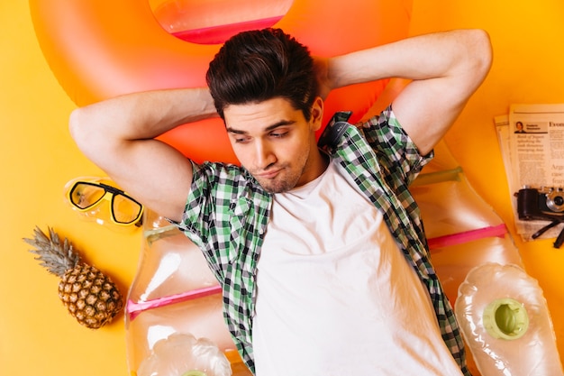 Free photo brunette man is lying on inflatable mattress on orange space with pineapple, dive mask and newspaper.