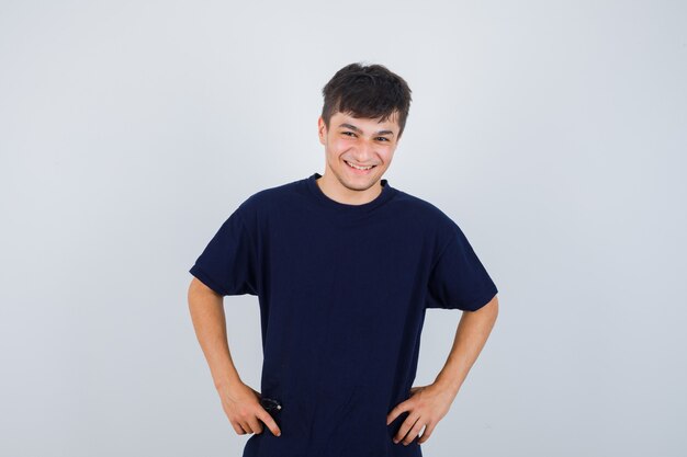 Brunette man looking at camera, holds hands on waist in dark t-shirt and looking happy , front view.