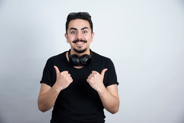 Brunette man model standing in headphones and showing thumbs up against white wall . 