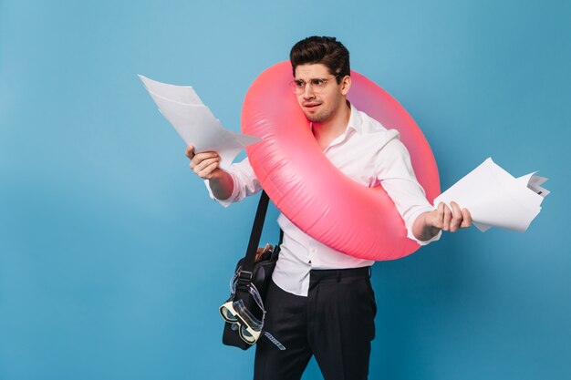 オフィスの服を着たブルネットの男は、不快感を持って文書を見ています。男は旅に出て、ピンクのインフレータブルサークルでポーズをとっています。