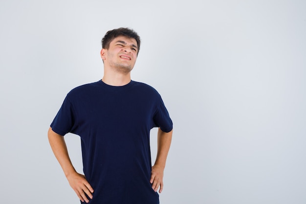 Brunette man in t-shirt holding hands on waist and looking painful , front view.