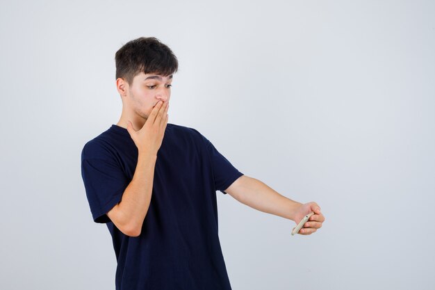 Brunette man in t-shirt looking at mobile phone while holding hand on mouth and looking perplexed , front view.