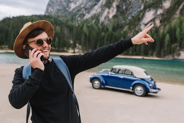 Brunette man wears sunglasses and black sweatshirt speaking on phone during walk along river shore and pointing finger on something interesting