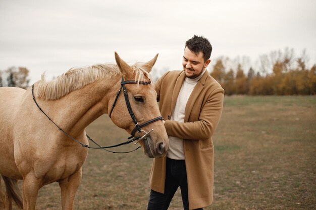 Brunette man with beard and brown horse standing in the field. Man wearing beige coat. Man touching the horse.