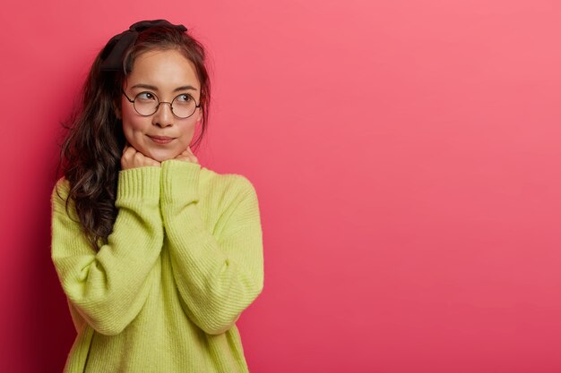 Brunette millennial young Asian woman keeps hands under chin, wears transparent glasses