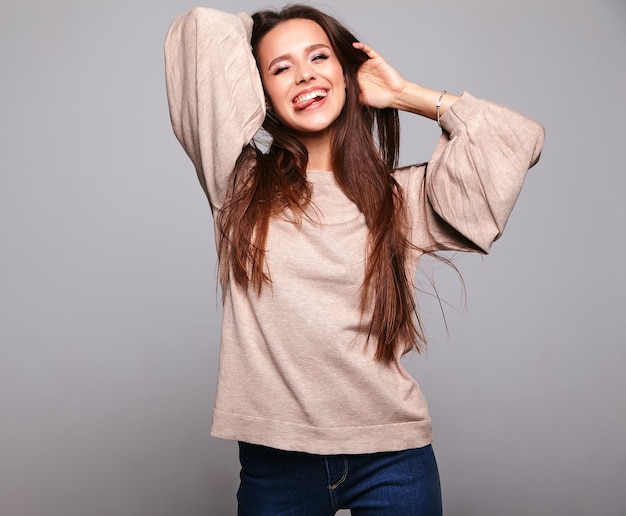 брюнетка в повседневной одежде бежевого теплого свитера на сером показывает язык