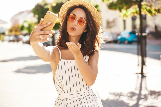 공기 키스를주는 휴대 전화를 사용하여 거리에서 포즈 여름 옷을 입고 갈색 머리 모델