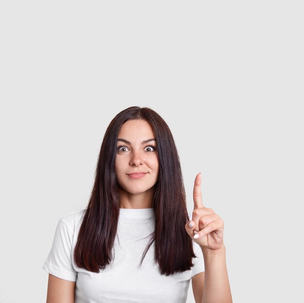 brunette mysterious woman with long dark hair, points with index finger upwards, has surprised expression